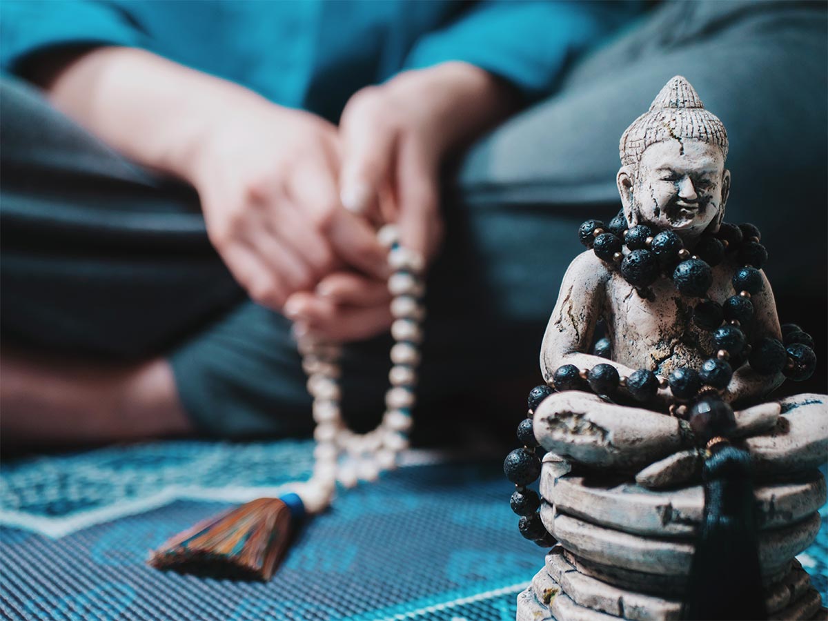 white hands hold a necklace while a person lays on a mat or carpet in prayer next to a statue of Buddha.