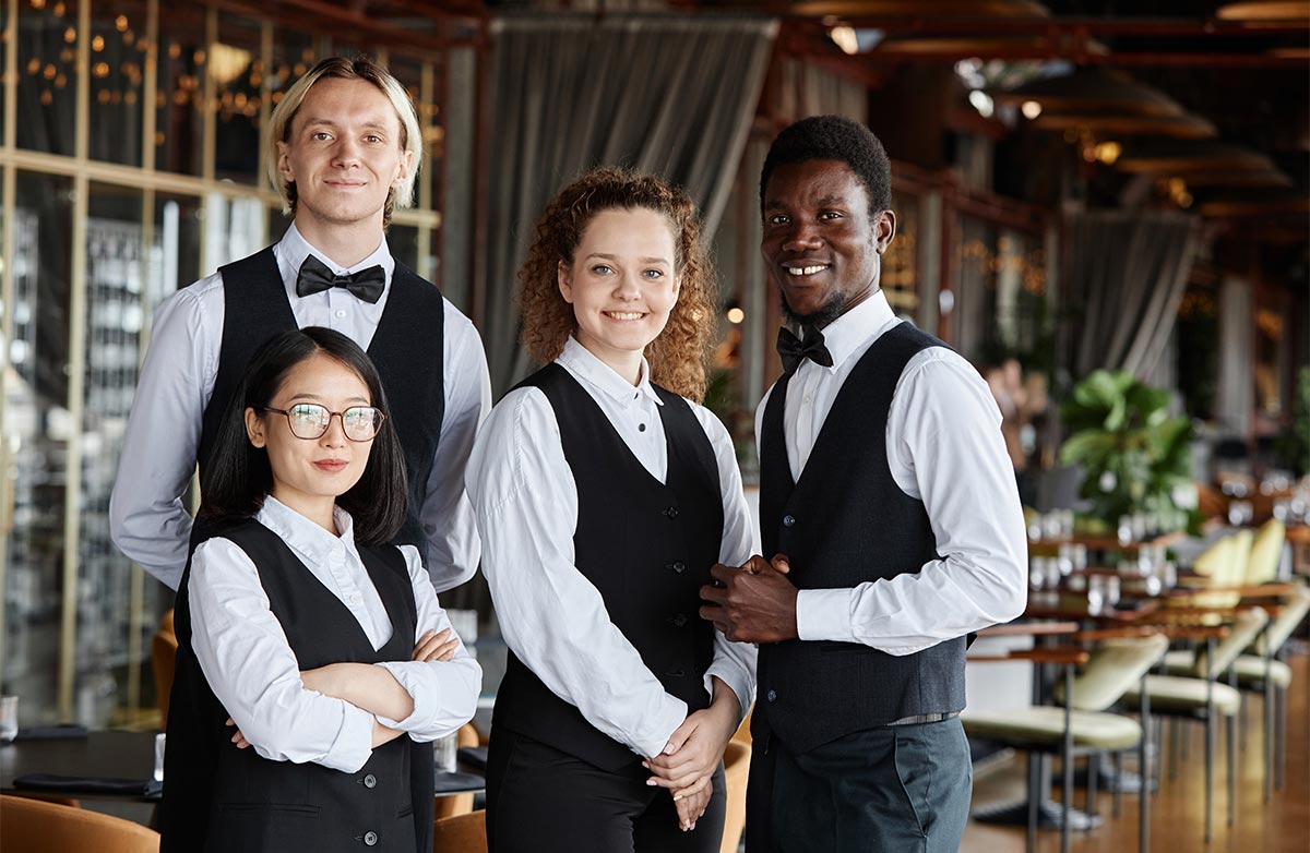 Four multiracial adults in their 30s dressed in classic server tuxedos smile at the camera in a nice restaurant