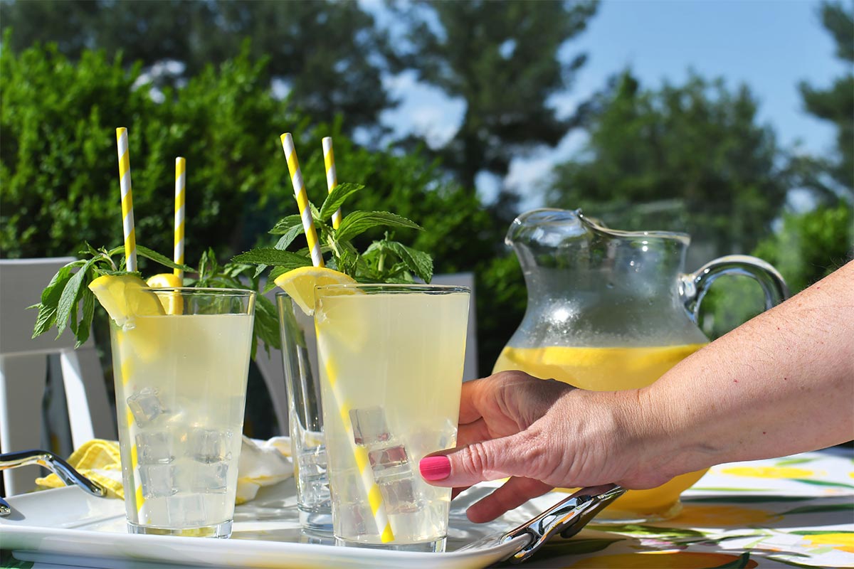 older white woman with pink nail polish reaches out for a glass of lemonade that sits on a tray next to two other glasses and a pitcher of lemonade on a sunny day