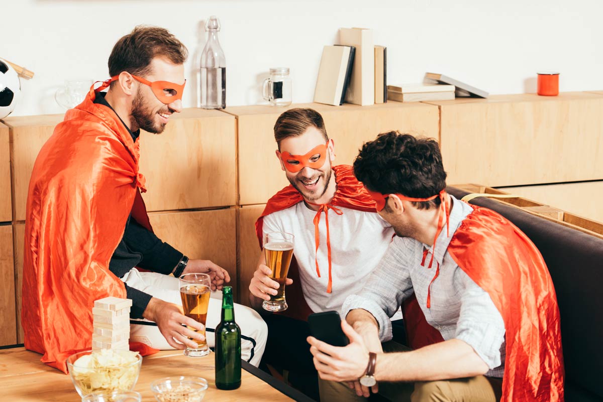 3 20 somethings white guys dressed in superhero costumes sitting at a bench and drinking beer in a circle, while smiling at each other. 