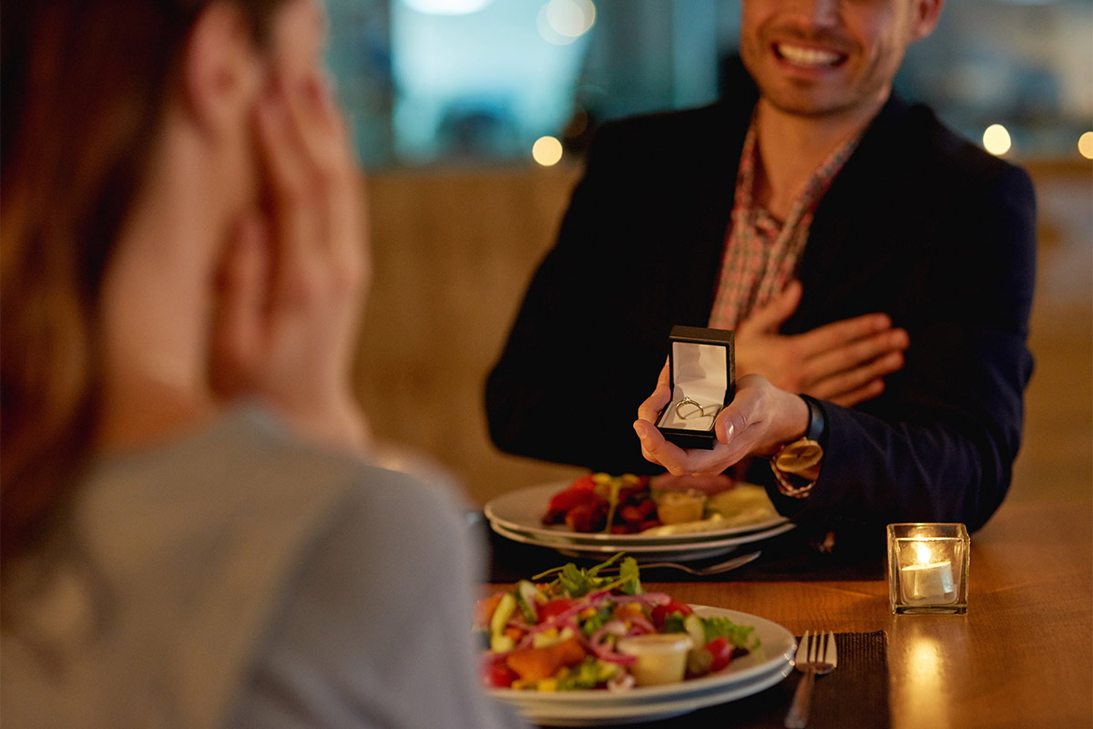 man joyfully presenting a ring in a box at dinner, smiling with a hand over his chest across the table as the woman reacts in surprise, hands on her cheeks