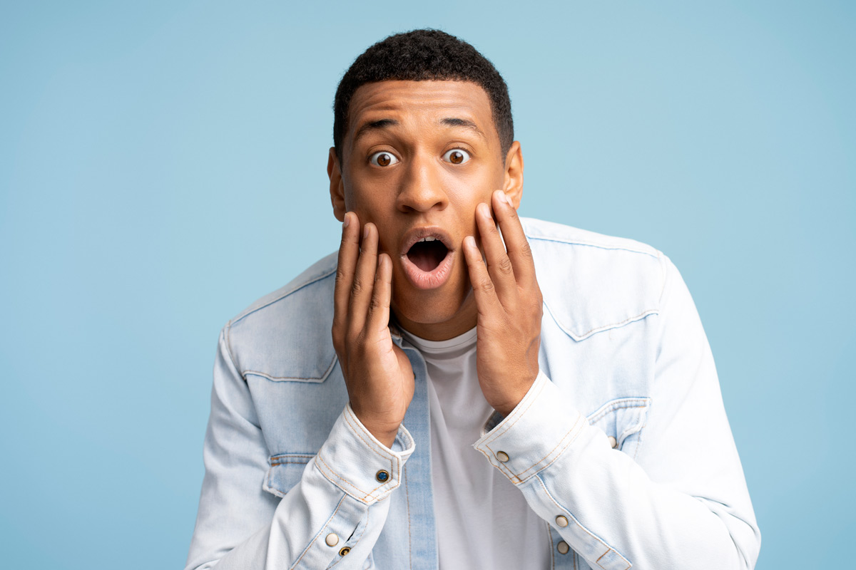 Black 26-year-old man looking shocked with his hands on his face in front of a blue background. 