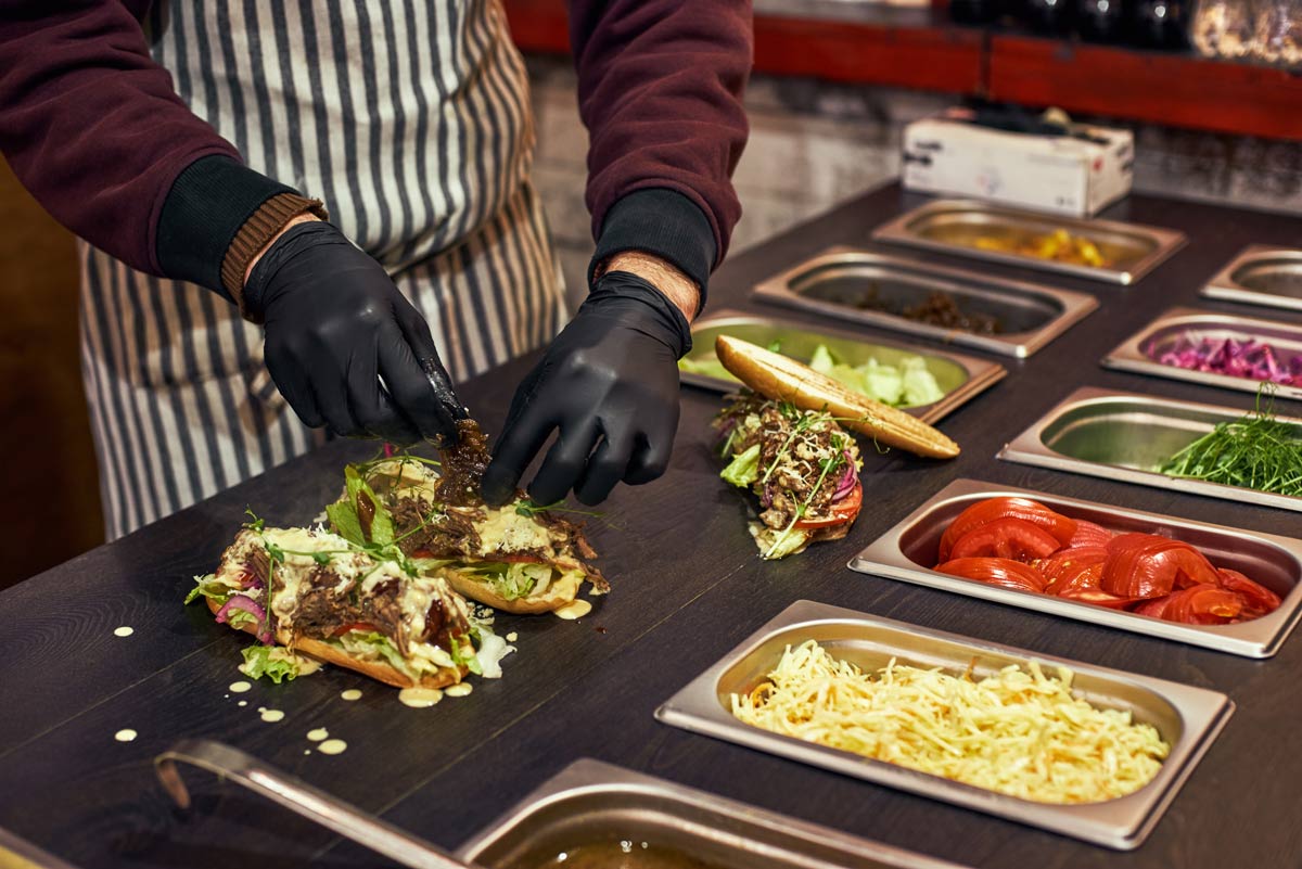 Person wearing gloves and a apron putting together tacos with taco ingredients in containers on a table in front of them