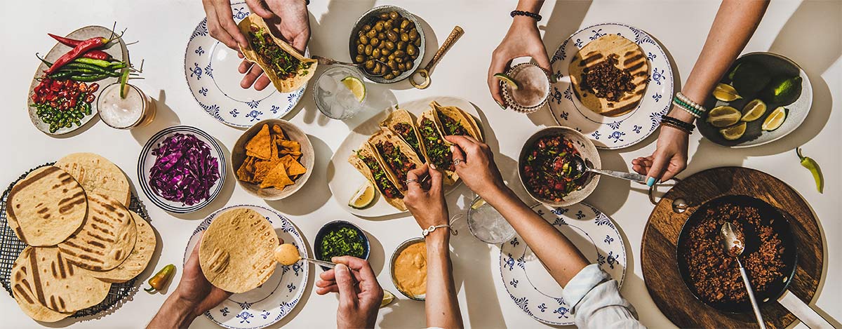 four people making tacos for dinner with tortillas, beef, limes, queso, chips, peppers, and other ingredients in bowls and plates spread out across the long white table