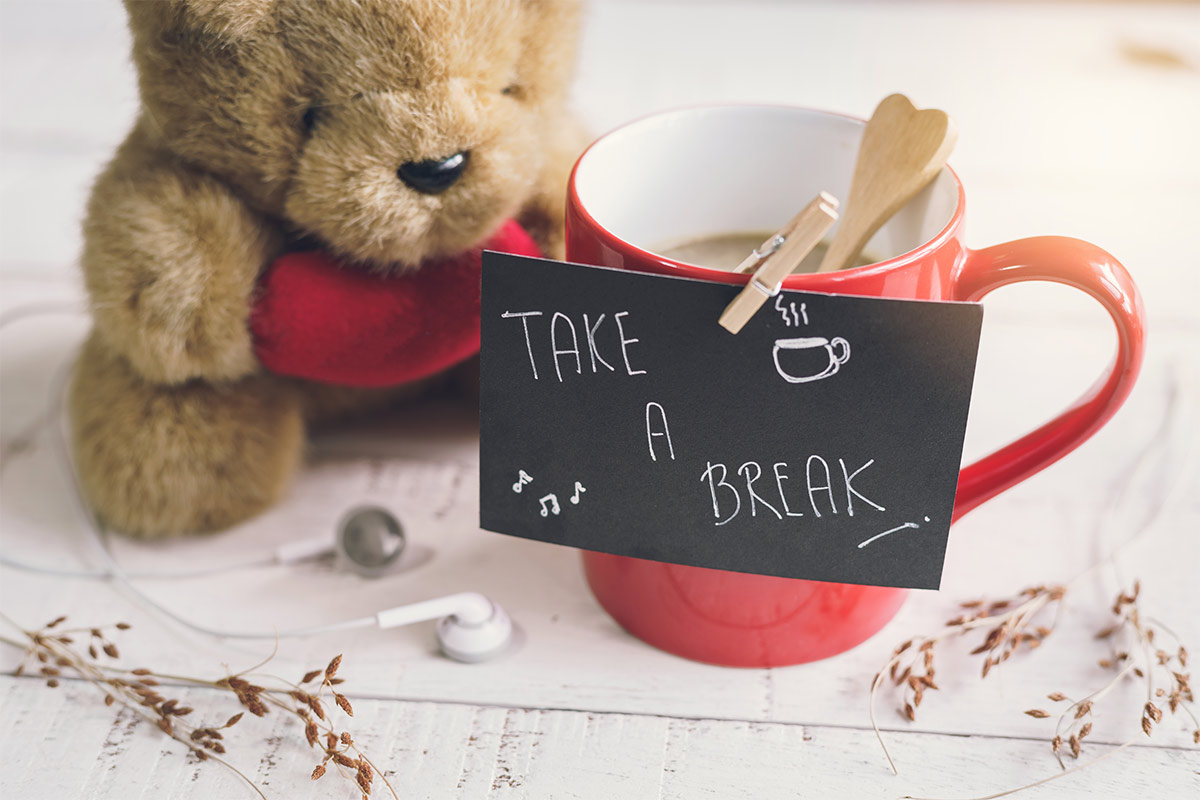 Brown teddy bear holding a red heart and a red cup of coffee on a table with a note attached to it saying ‘Take A Break’, symbolizing the importance of taking breaks to de-stress before your wedding.