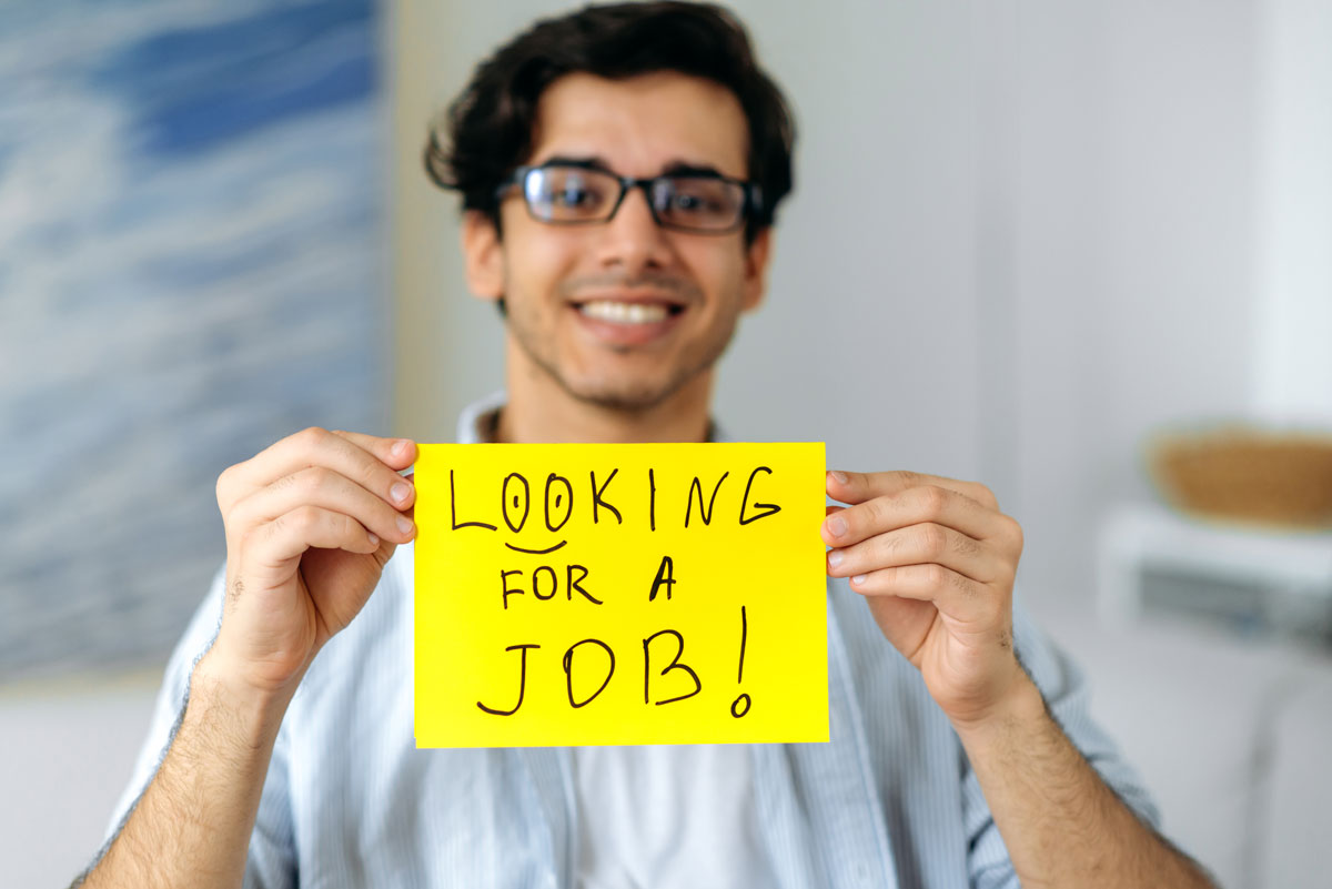 White man in his 30s with glasses holding a bright yellow sign saying “Looking for a Job!”.