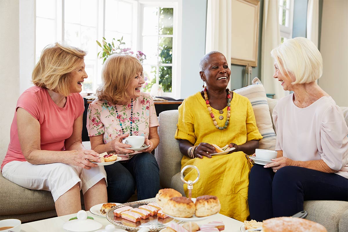  Four multiracial women in their 60s smile and laugh as they sit on a couch holding tea and pastries next to a coffee table of assorted pastries 