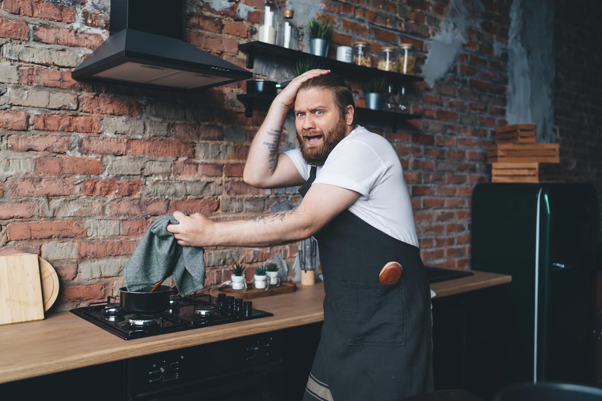 30-something distressed white man with a hand on his head and holding a cloth above a stove, in a kitchen