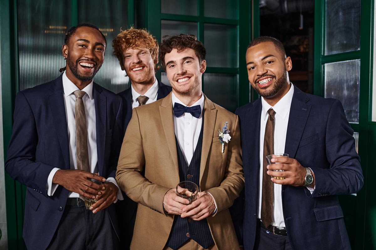 Mixed-raced 20 something groomsmen with a white groom in the middle of them holding glasses filled with wine. 