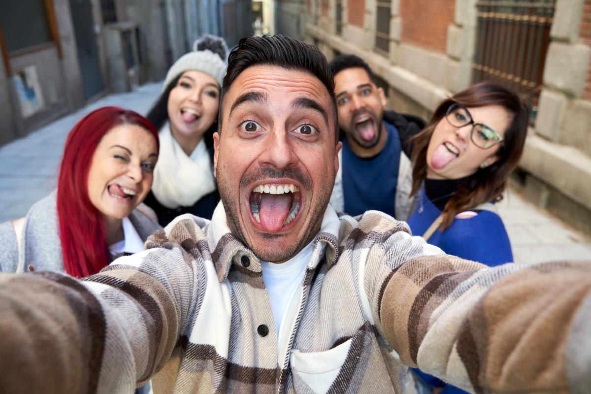 latino man snapping a silly selfie with four friends, everyone sticking out their tongues and laughing outside