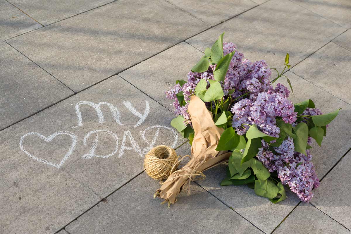 purple flower bouquet in craft paper and twine on concrete tile next to the phrase “my dad” and a heart in chalk