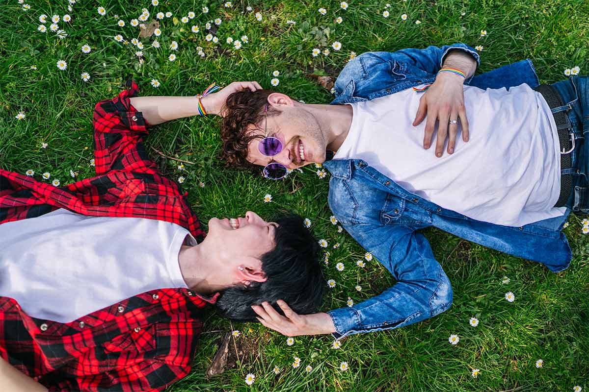 Gay multiracial couple lays on grass surrounded by flowers smiling and playing with eachothers hair