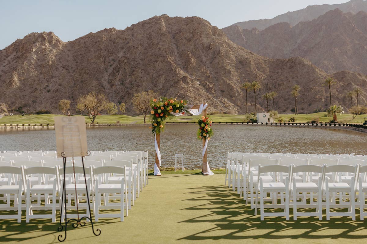 Wooden altar with white linen and bright flower pieces sits in front of white chairs with mountains and a pond behind it