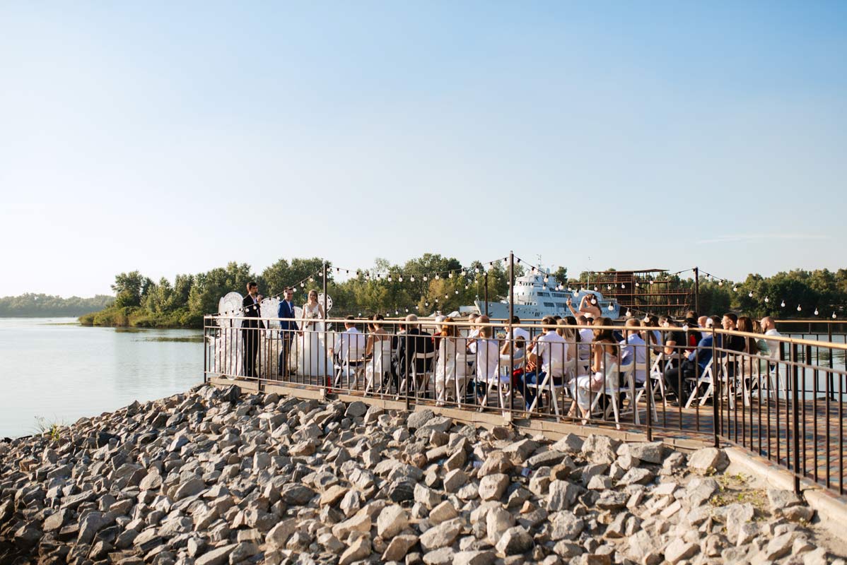 Far away shot of an outdoor wedding ceremony taking place on a wooden dock overlooking the water during a sunny day 