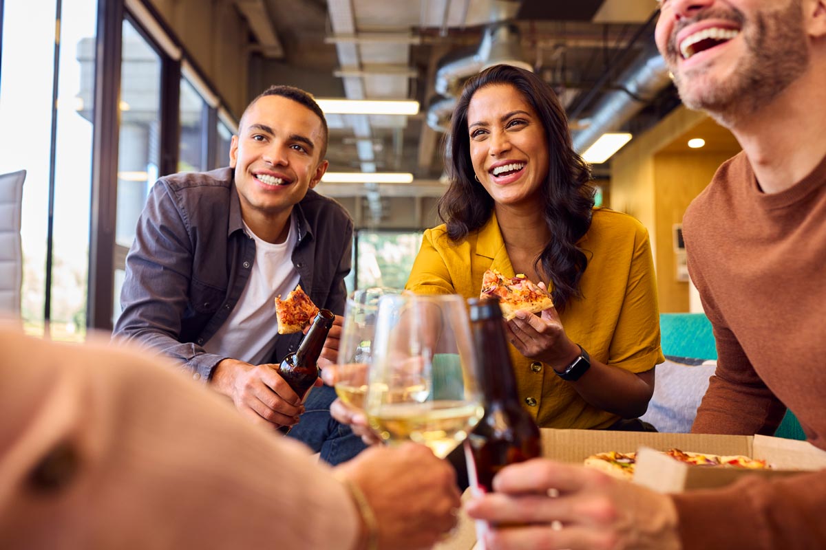  A group of 4 white coworkers in their 30s and 40s share wine and pizza, while they are all smiling.  