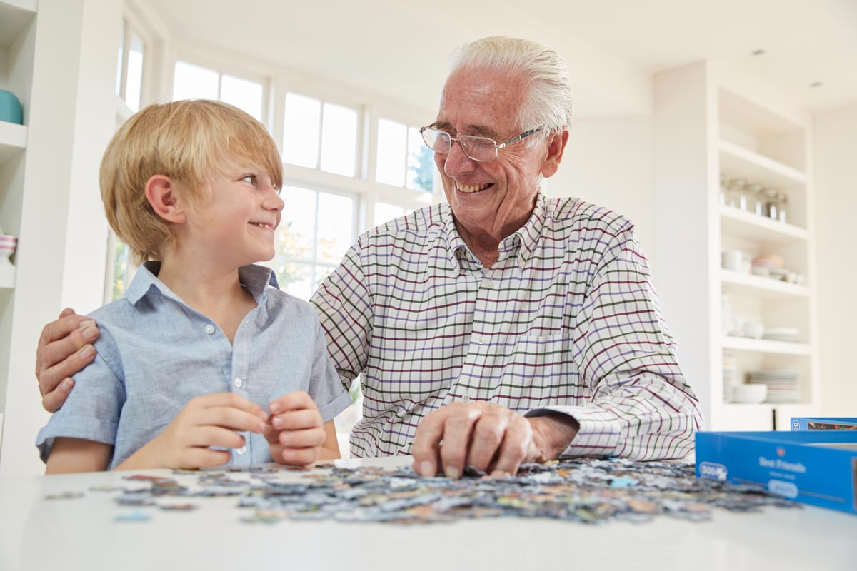 4-year-old white grandson doing a puzzle with his 70-year-old grandfather wearing glasses inside their house smiling at each other