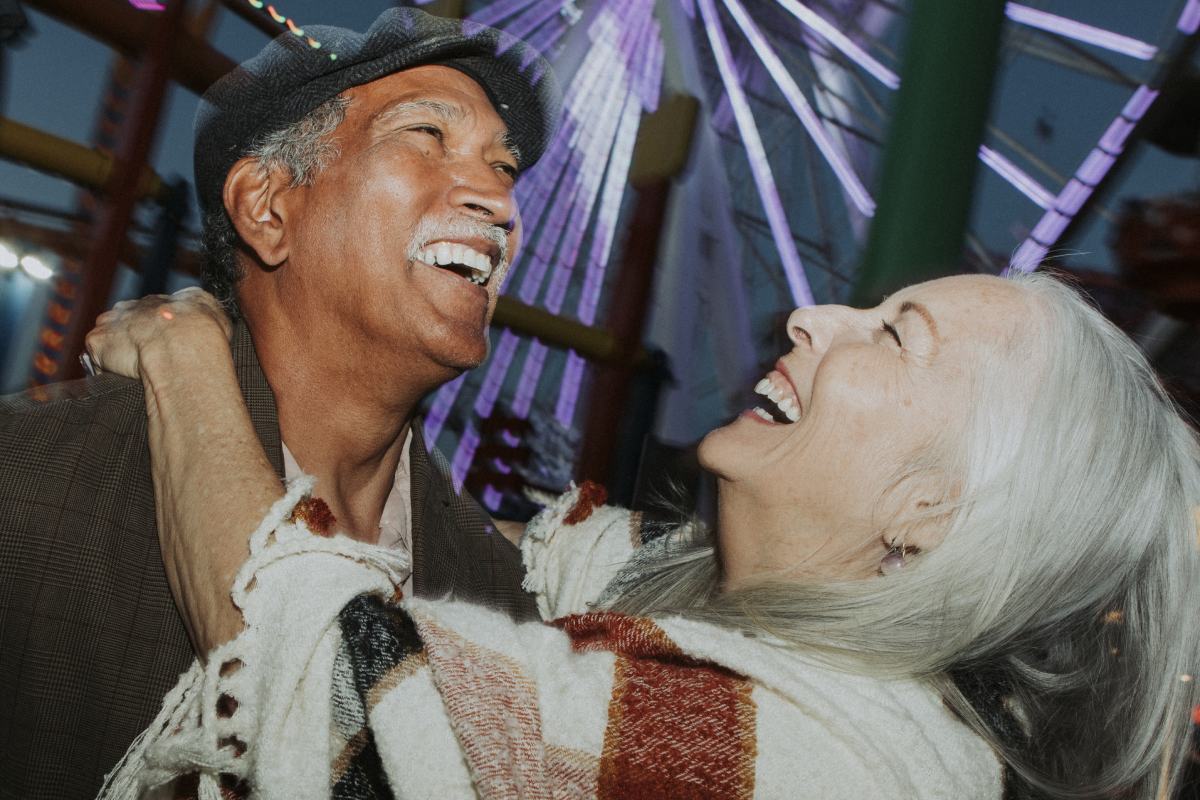 black elderly male laughing with white elderly wife with her arms around him in front of a Ferris wheel 