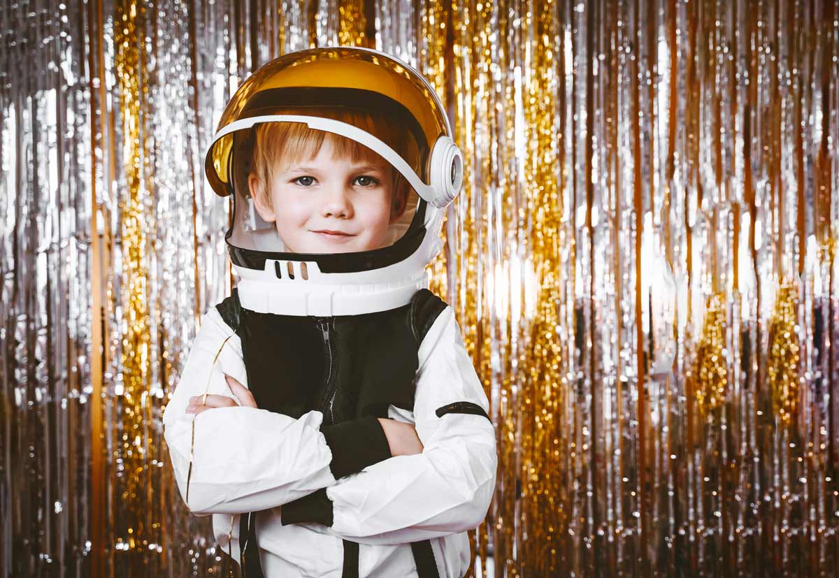 A young white boy dressed as an astronaut crossing his arms in front of a gold and silver background.