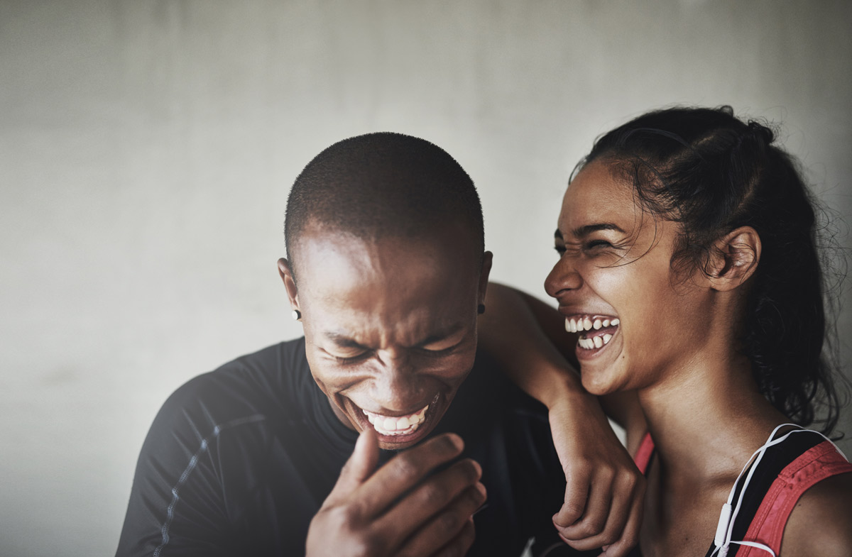 a 20 something black couple laugh uncontrollably after a workout while the woman lays her arm on her partner's shoulder.