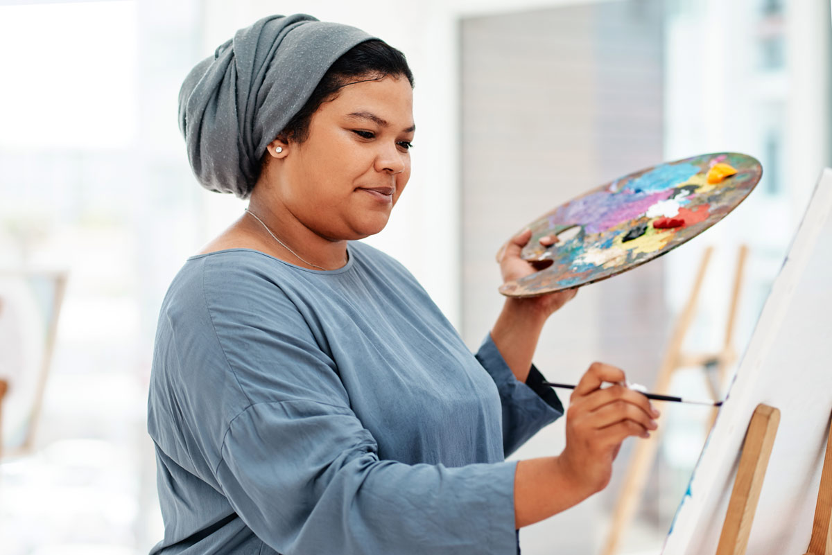 Black 40-something woman in hair turban holds a paint palette as she uses a paint brush to paint on a canvas