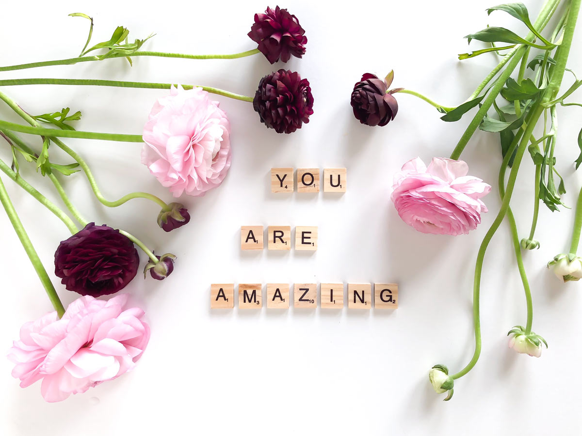 Letter tiles spelling “you are amazing” laid out on a white table surrounded by pink, white, and dark purple flowers.