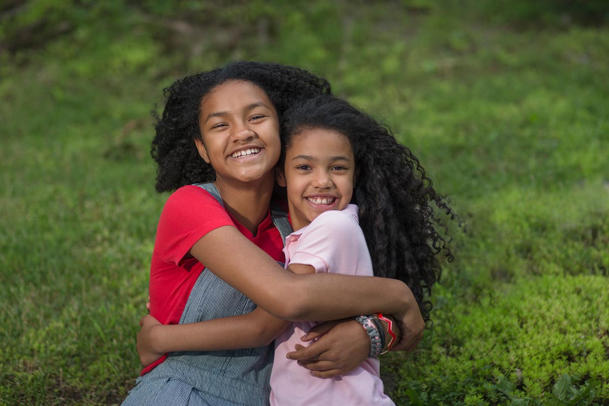 Two black sisters age 12 and 8 smile and hug each other from the side while sitting in the grass 