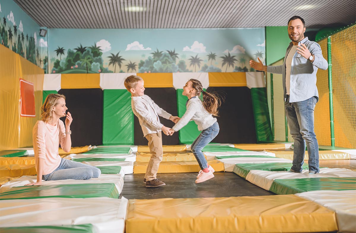 two young white children jumping and holding hands on a trampoline at a trampoline park as white 30 something woman and man watch them