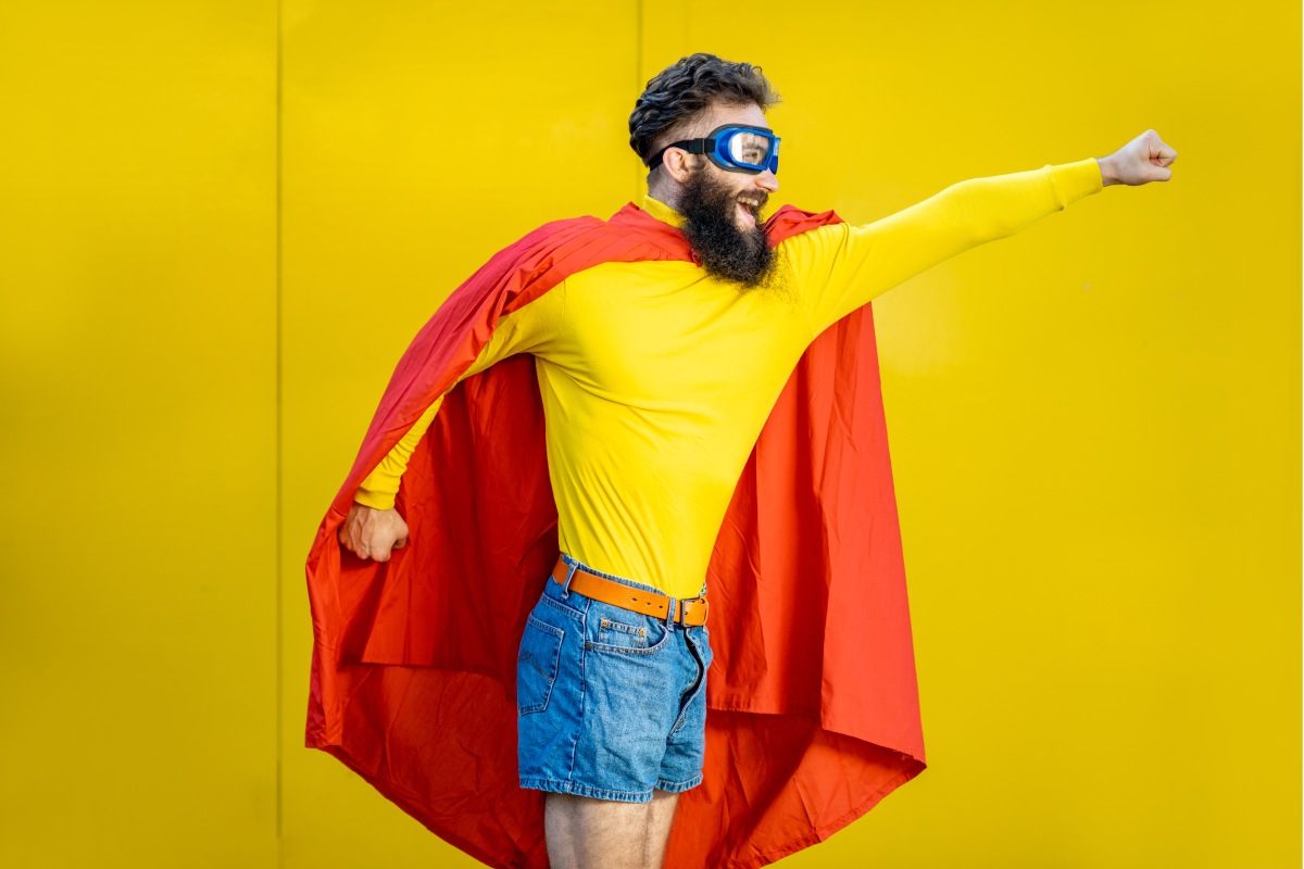 40 something white man with dark beard dressed as superhero doing a superhero pose and smiling against a yellow background 