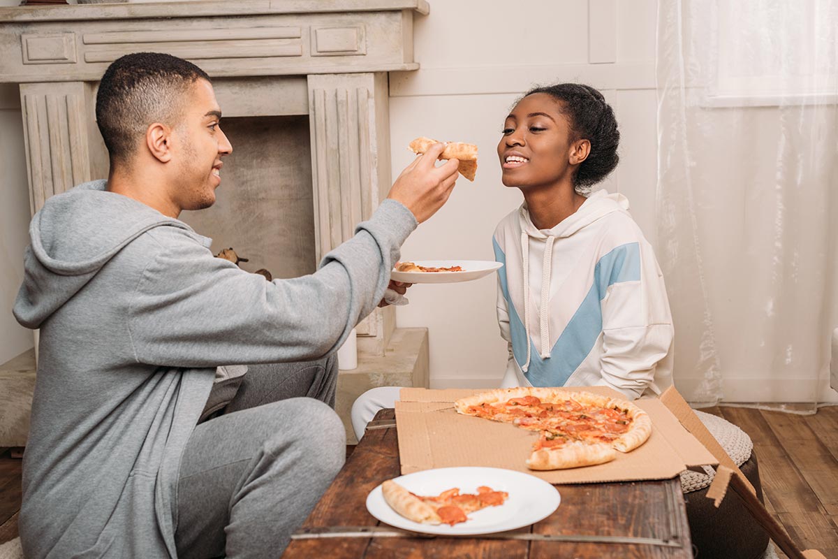 20 something black man feeds 20 something black woman pizza while sitting on floor by fireplace in sweatshirts