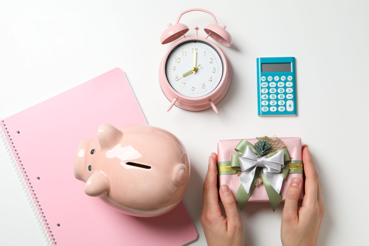 Person holding a small pink wrapped gift box with a green, gold, and silver bow, and a pink piggy bank, pink and gold analog clock, and blue and white calculator