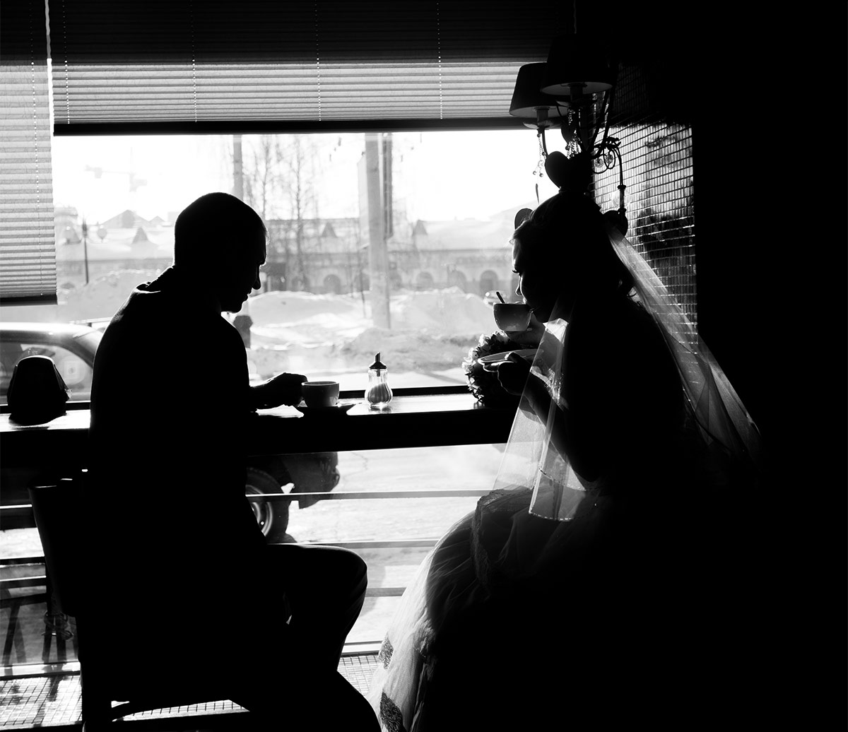  Black and white silhouette side profile of a wedding couple drinking coffee at a hightop table by a window in a coffee shop