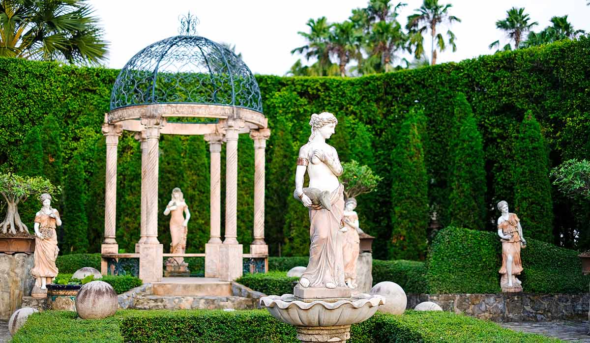 Historical garden with statues and a dome-like structure in the middle surrounded by tall manicured hedges and palm trees in the background. 