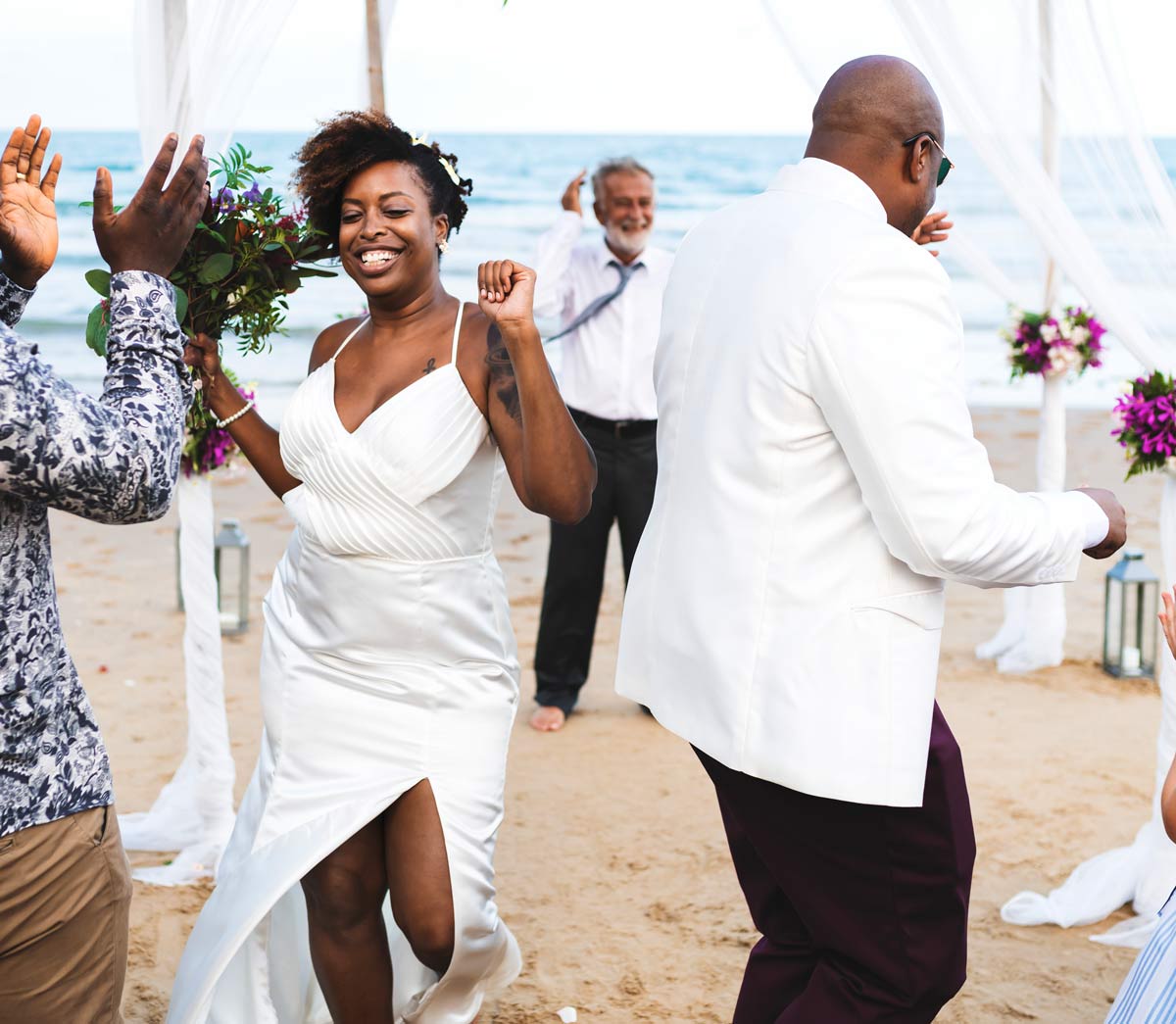 black bride holding bouquet and joyfully dancing with her father on the beach with guests clapping