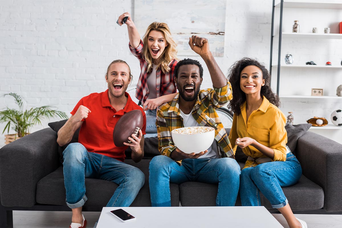 Multiracial group of young adult friends in their 30s-40s cheer on couch at home holding white popcorn bowl and an American football 