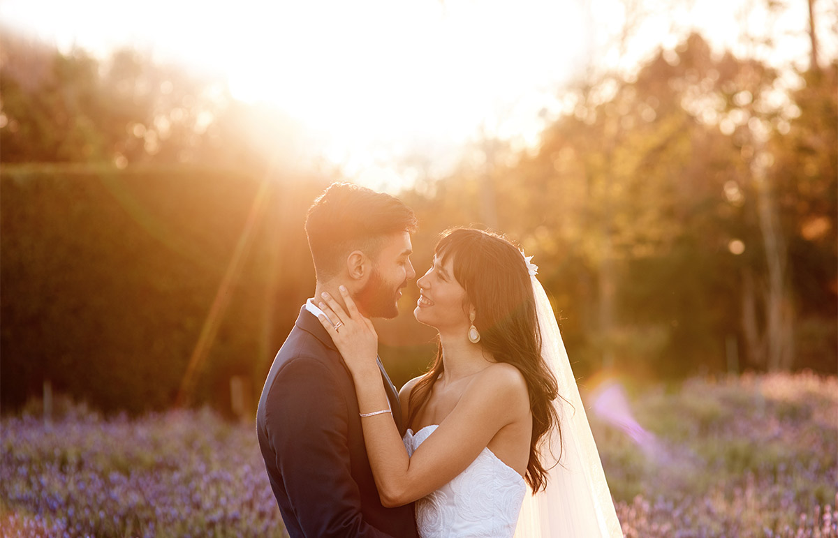 Early 30s bride and groom stand in a field of purple wildflowers during golden hour with the sun lighting up the background.