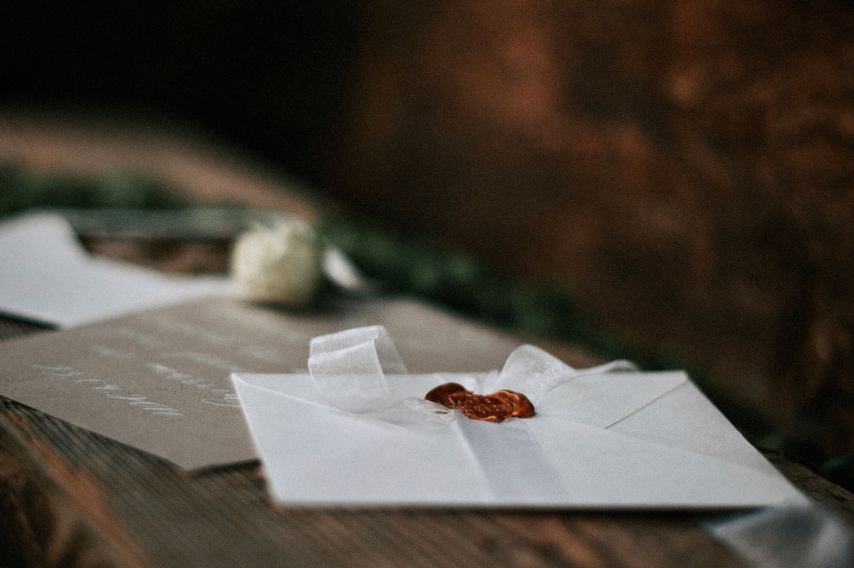 Brown wedding invitation and white envelope with a red wax seal and white bow sitting on a wood table.