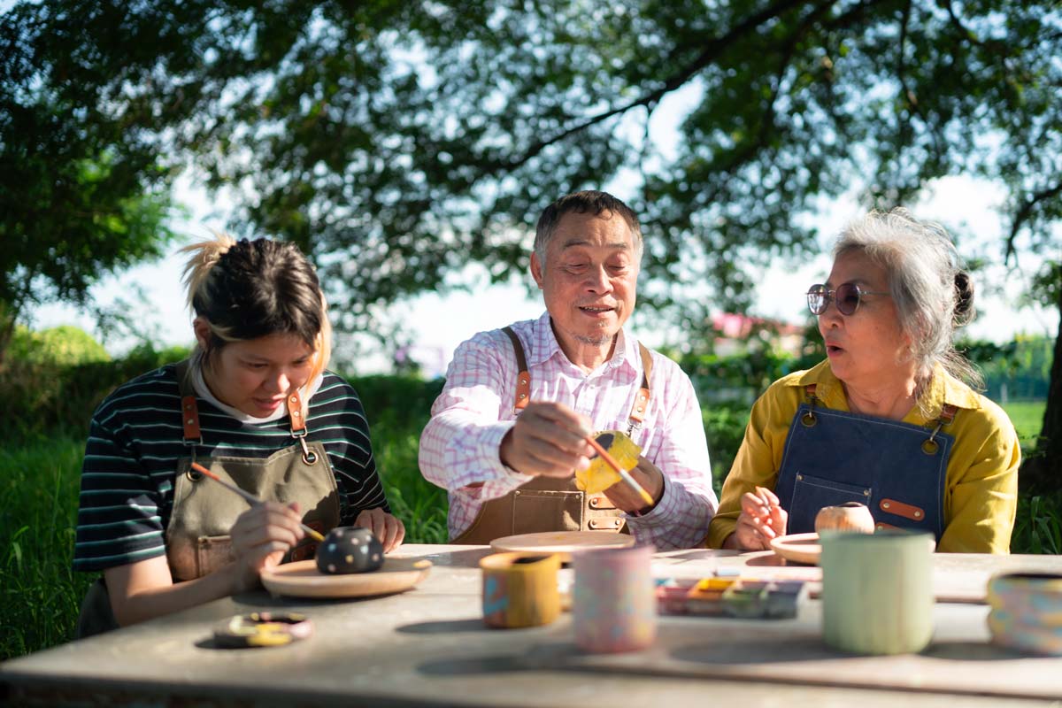 Two asian women and an asian man engaged in pottery making at an outdoor crafting station.