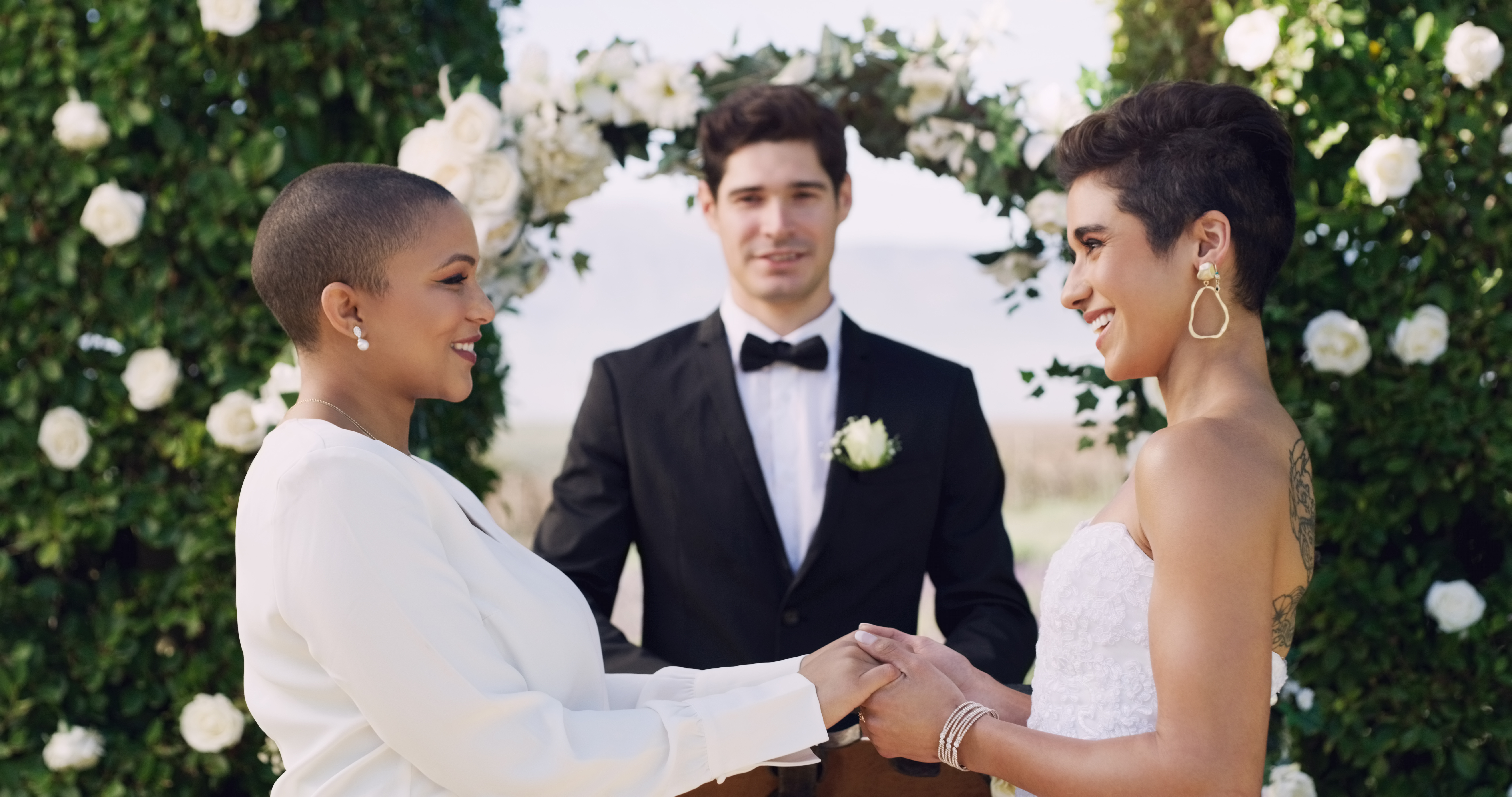 20 Somethings lesbian Black women couple wearing white and holding hands and looking at each other while standing at the altar surrounded by green shrubs with white roses and a 20 somethings white man standing in between them wearing a suit.