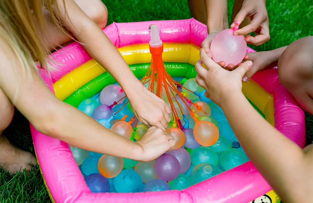 White little kids hands grabbing rainbow colored water balloons in a kitty pool, while they kneel on the grass outside. 