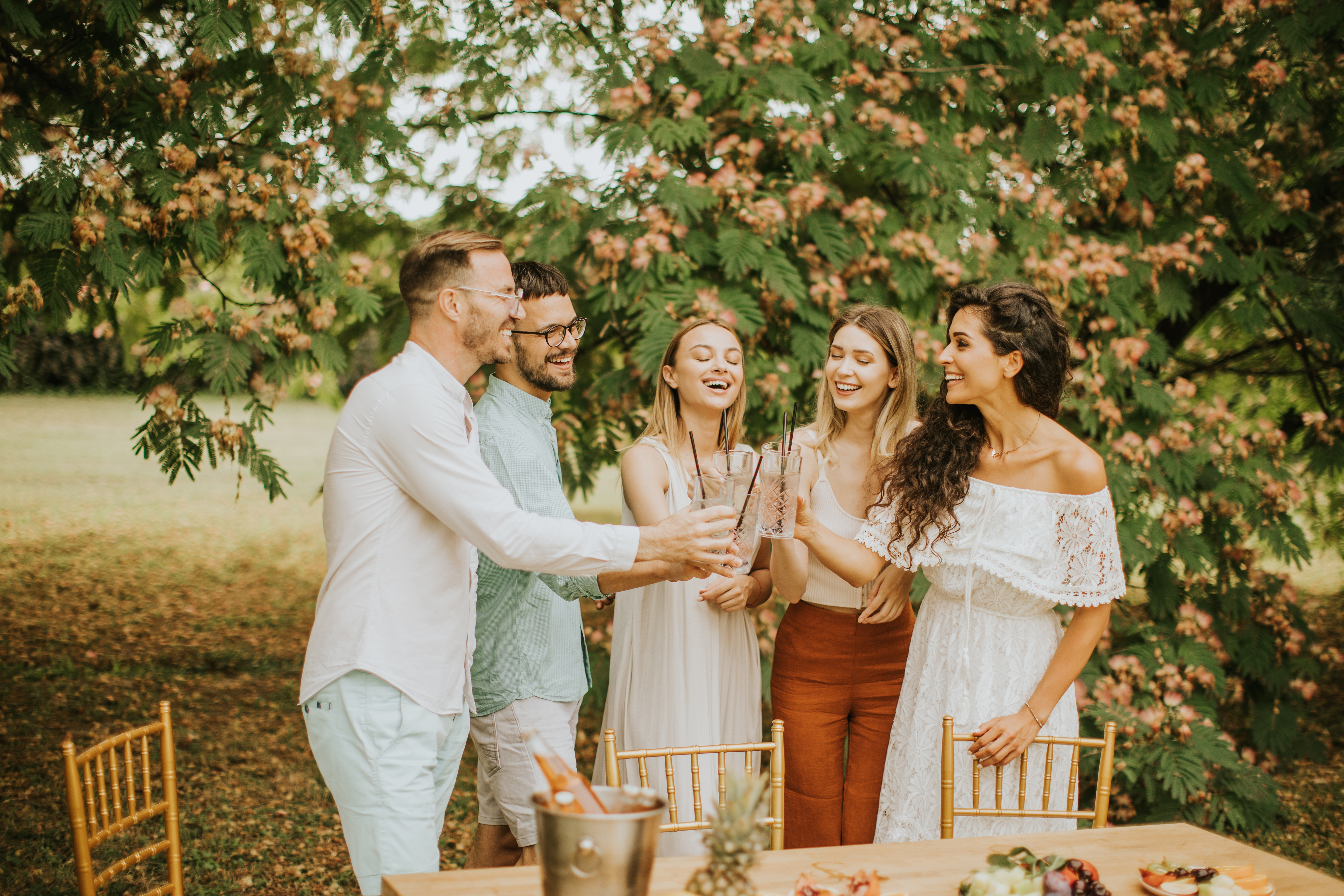  20 somethings white men and women friend group wearing elegant clothes, grouped in a circle clinking glasses together and smiling while being outside next to a table.