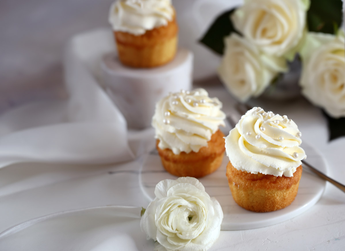 Vanilla cupcakes with white frosting sit on a white table with white roses