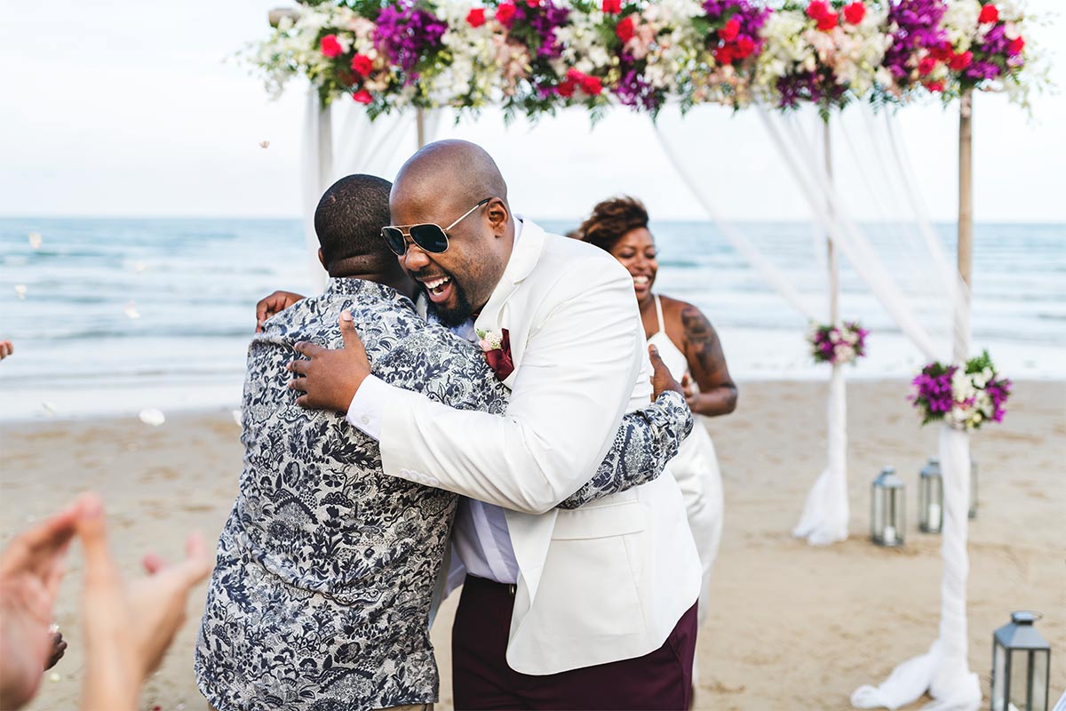 Black 40-something groom wearing white suit jacket and sunglasses smiles and hugs black man in paisley long sleeve as they stand in the sand by a colorful, floral decorated wedding arch on the beach during a sunny day