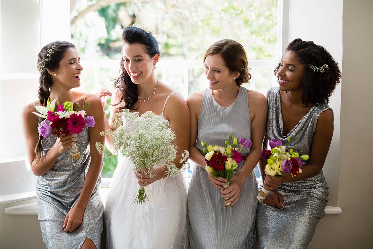 Three multiracial bridesmaids in their early 30s wear light blue dresses and hold colorful bouquets as they laugh with a 30-something white bride holding a bouquet of baby’s breath as the sun shines through the window behind them