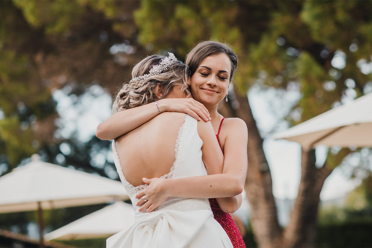 White 20-something brunette bridesmaid in red dress hugs white bride with jeweled headpiece as they stand outside near some trees during the daylight 