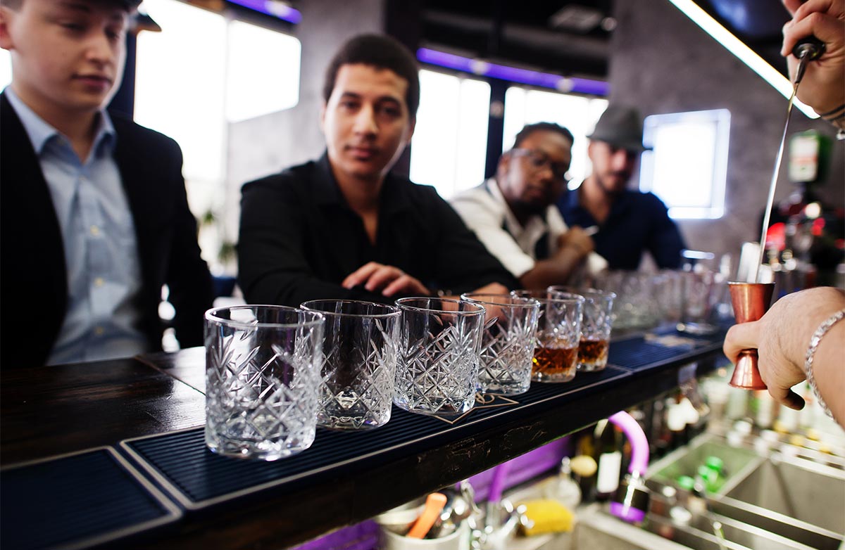 Line of whiskey glasses sit on a bar mat as the bartender’s hand pours a drink and four multiracial adult men sit at the bar behind them