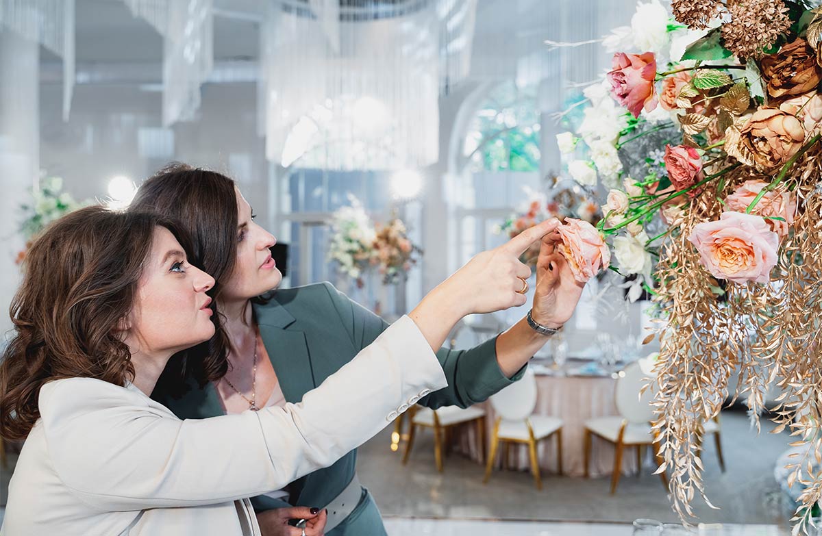 Two white women in their 40s wearing blazers point at and touch a large bouquet of assorted roses and gold detailing 