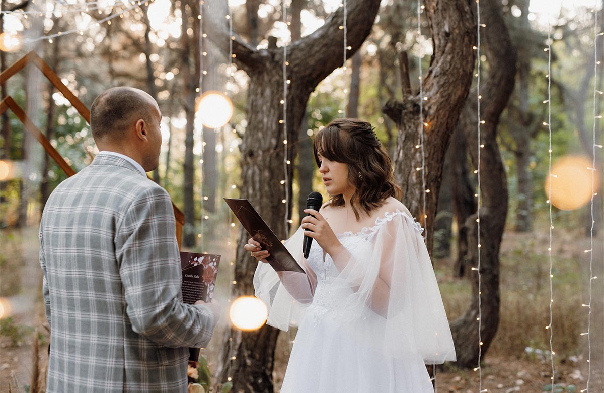 A white bride in her late 20s reads her vows on a microphone at an outdoor wedding surrounded by hanging twinkle lights