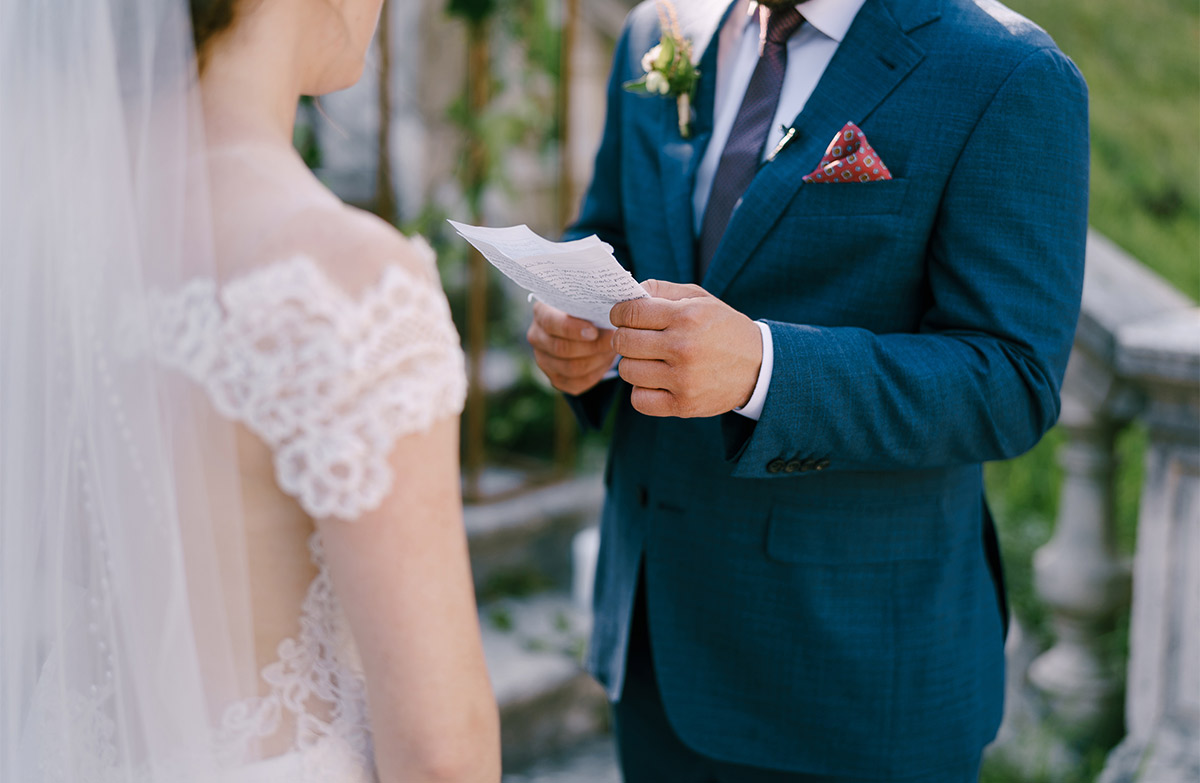 A white groom in navy suit reads his vows off a piece of paper to a bride.