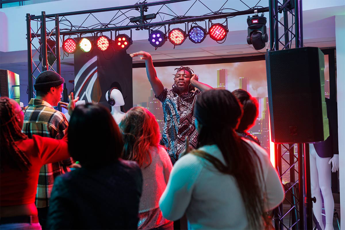 A black twenty-five year old DJ with his hand raised in the air, engaging with the energetic crowd before him, at a club.