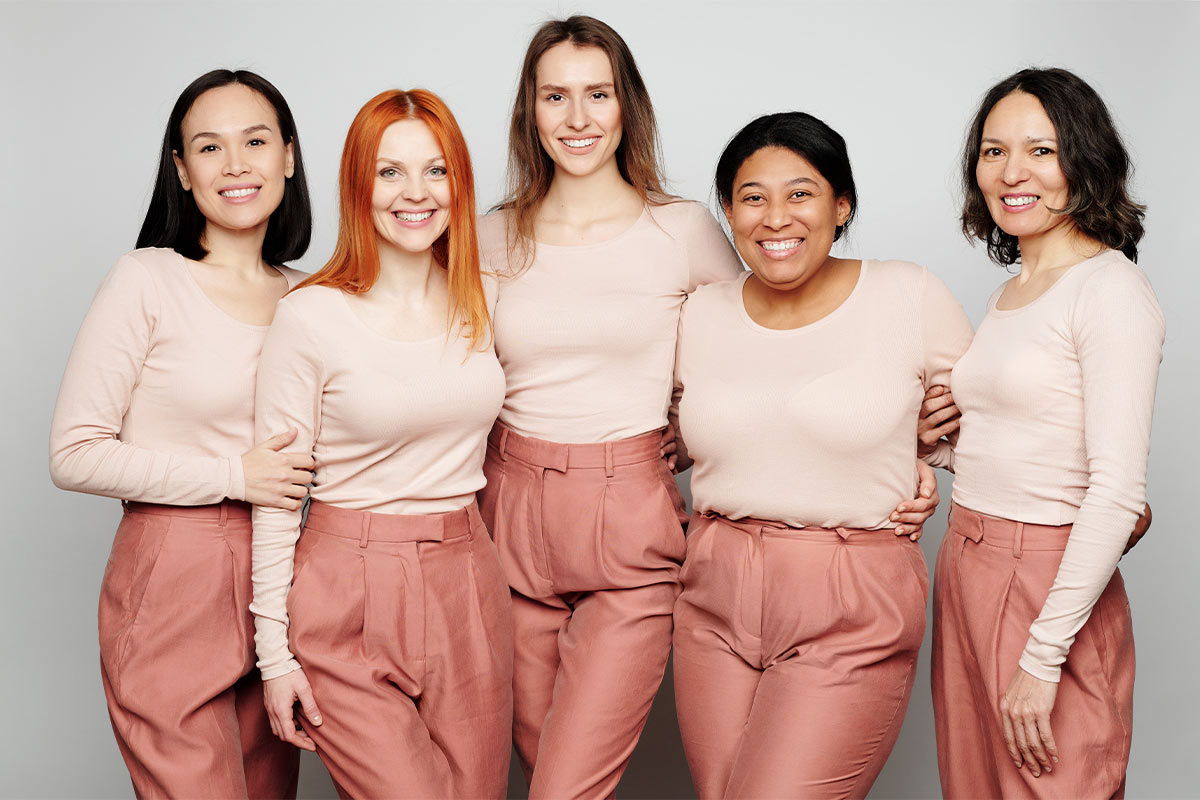 Five multiracial women in their 30s and 40s wearing matching nude long sleeves and mauve trousers stand and smile in front of a gray backdrop 